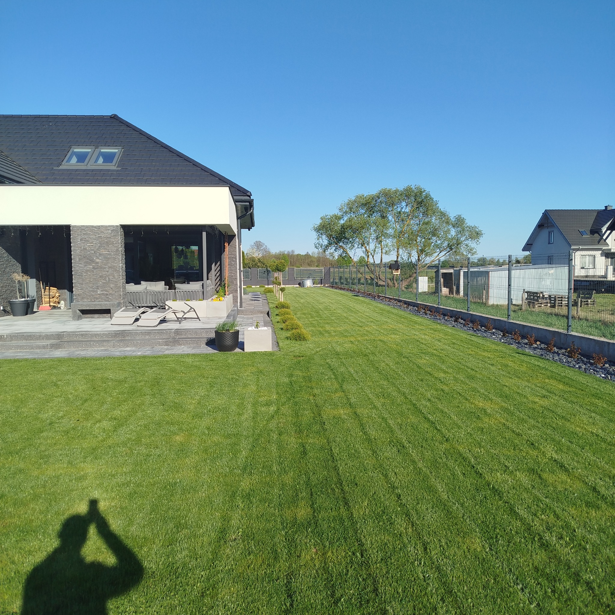 Nowoczesny ogród z idealnie przystrzyżonym trawnikiem, niskimi krzewami i ogrodzeniem panelowym. W tle dom z tarasem i meblami ogrodowymi.