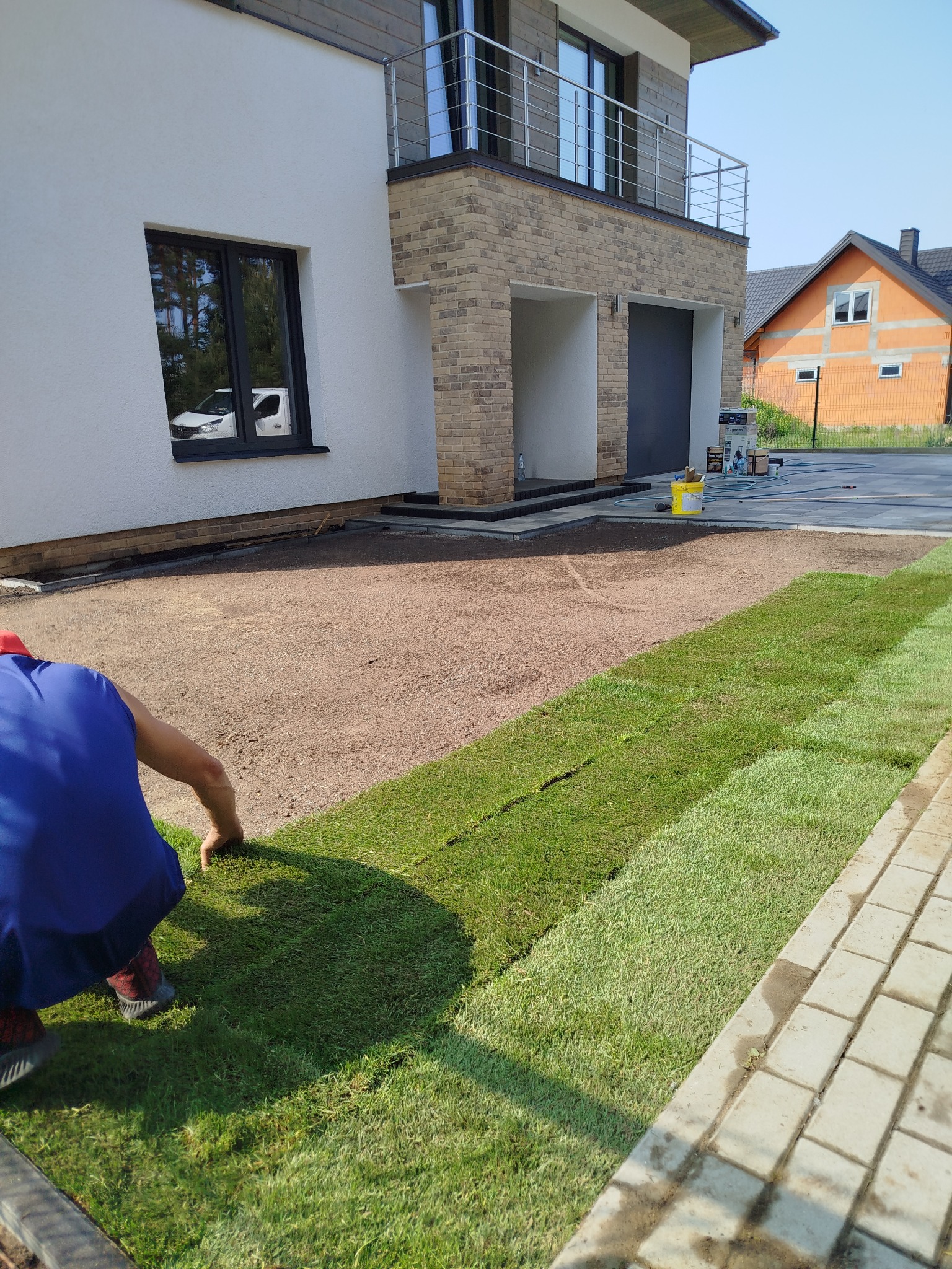 Układanie trawy z rolki na przygotowanym podłożu obok jasnej kostki. Widoczny fragment nowoczesnego domu z balkonem i sąsiedni budynek w tle.