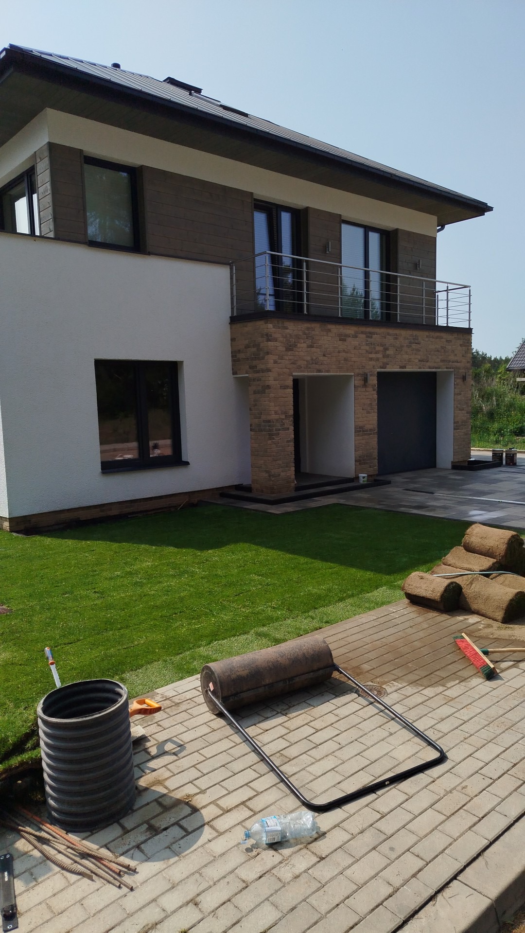 Nowoczesny dom z równo ułożoną trawą z rolki, wałem do trawy i paletą kostki brukowej. Widoczne detale jak ceglana elewacja i balkon.