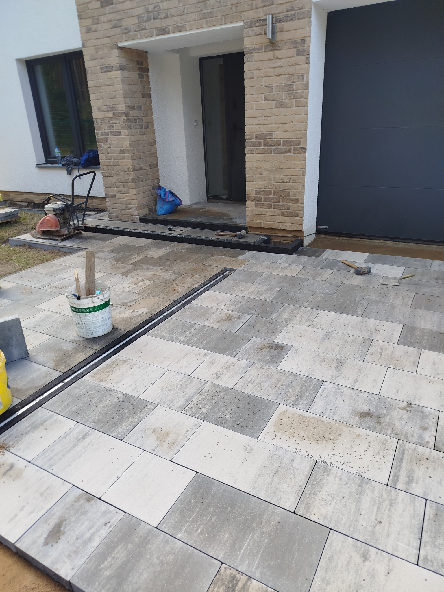 Układanie jasnoszarej kostki brukowej z odpływem liniowym przed budynkiem z beżową cegłą. Widoczny sprzęt budowlany i narzędzia pracy.