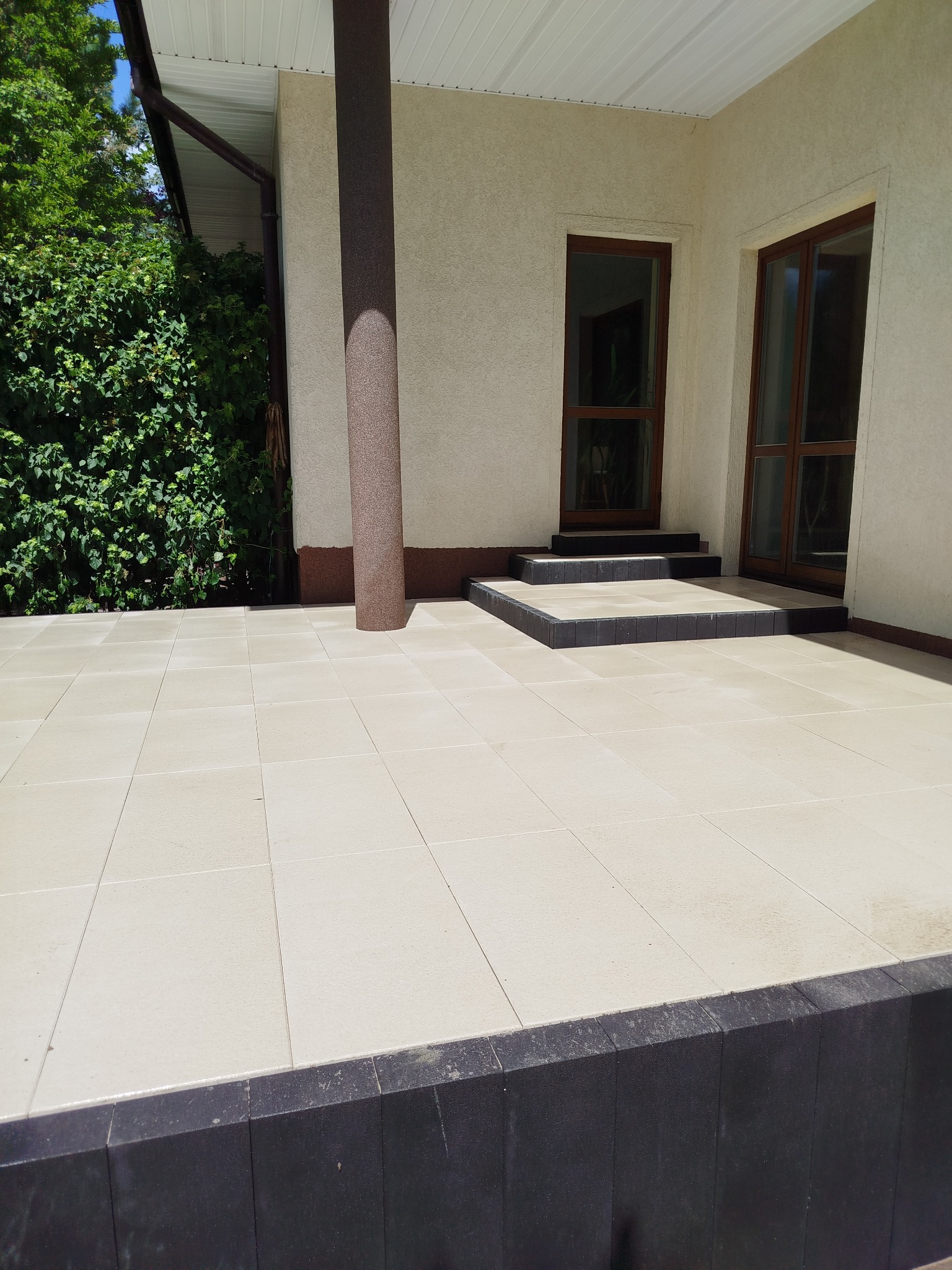 Jasna, kwadratowa kostka brukowa na tarasie domu z kolumną i schodami wejściowymi. Ciemne obrzeża kostki brukowej. Widok na zieleń ogrodu.