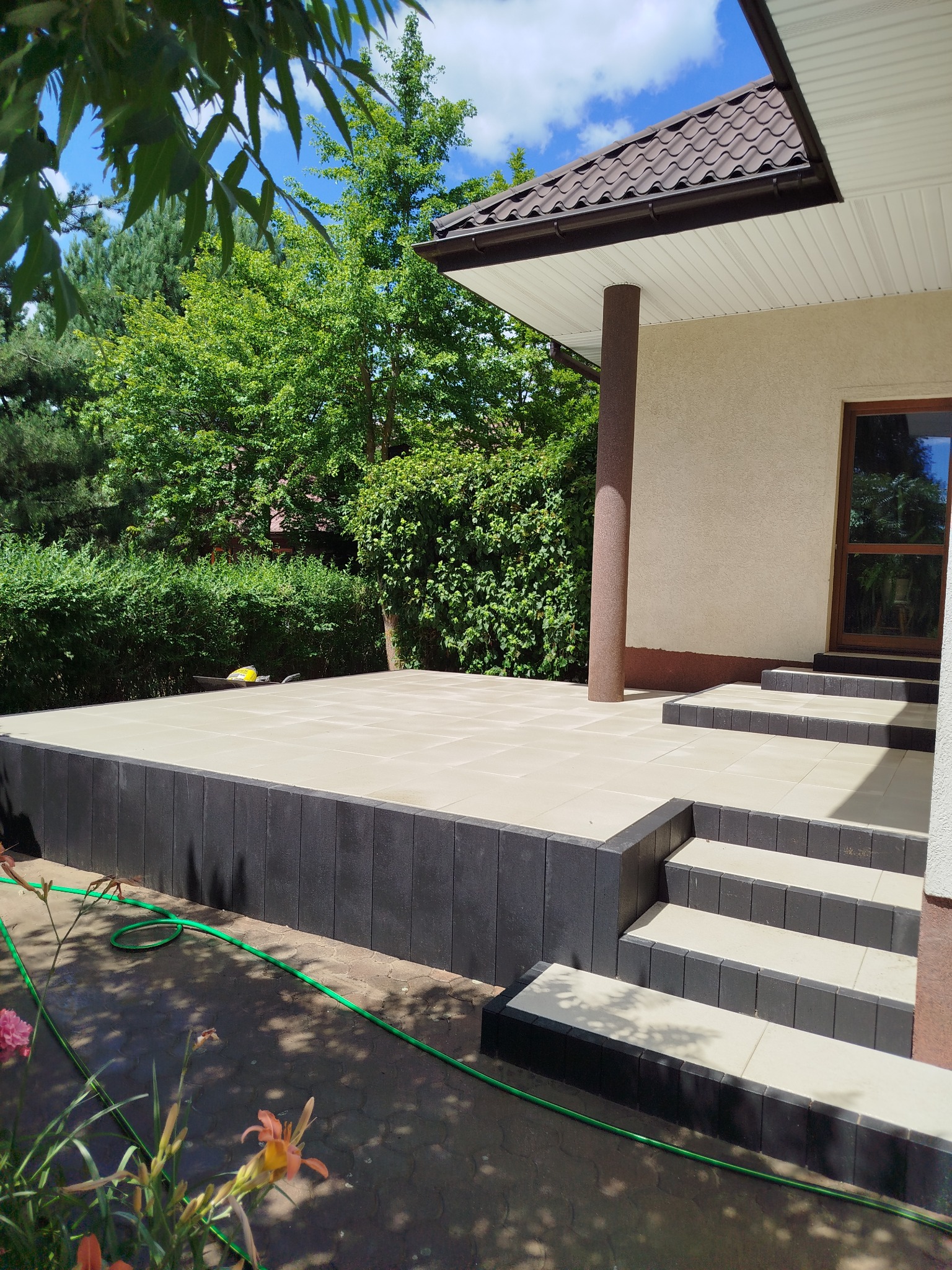 Nowoczesny taras z jasnych płyt i ciemnych obrzeży, schody wejściowe, zielony ogród w tle, słoneczny dzień. Widoczny wąż ogrodowy i fragment roślin.