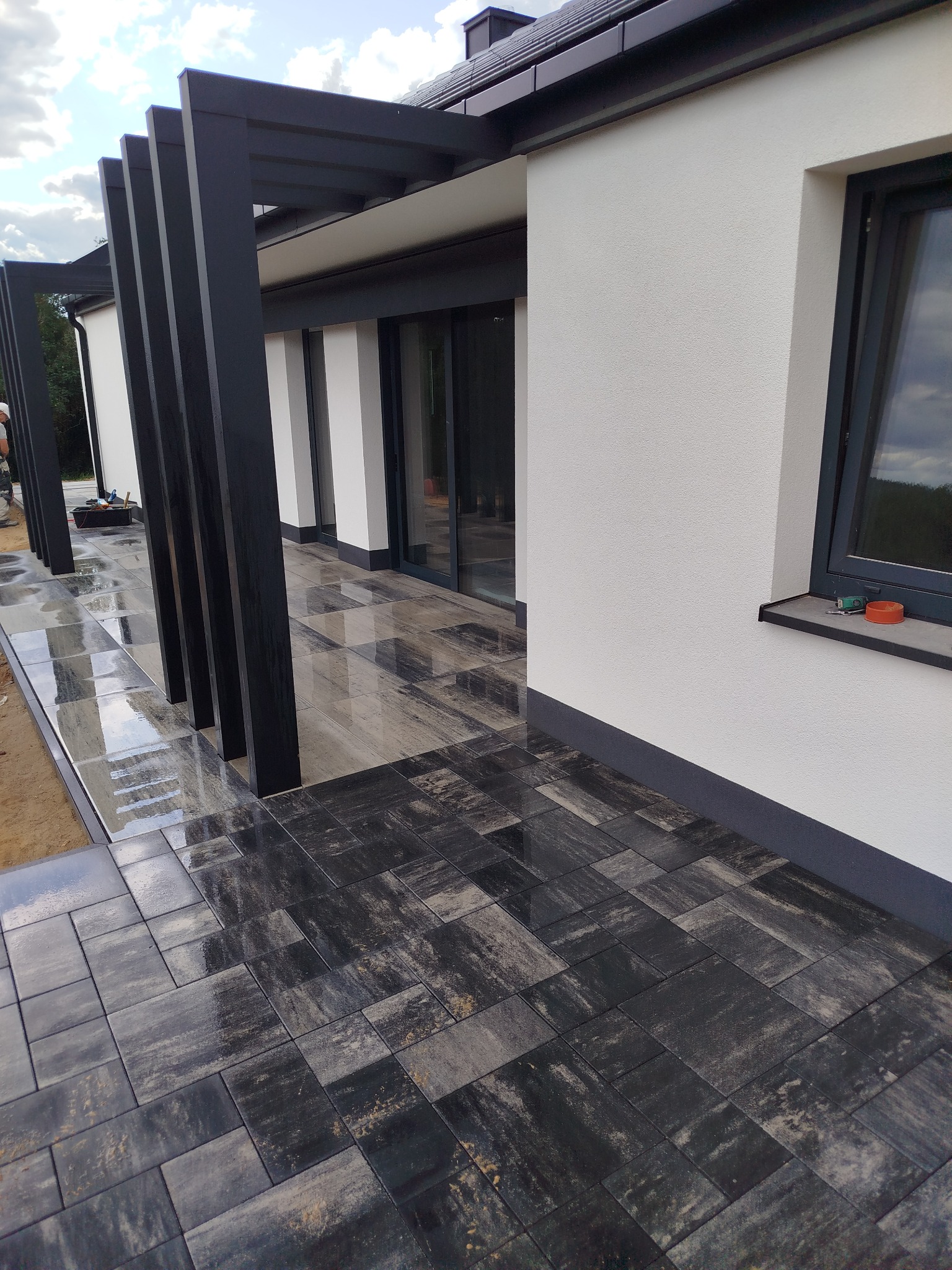 Nowoczesny taras z ciemnej kostki brukowej ułożonej przy białym budynku z czarną pergolą. Widoczne odbicia w mokrej nawierzchni kostki.