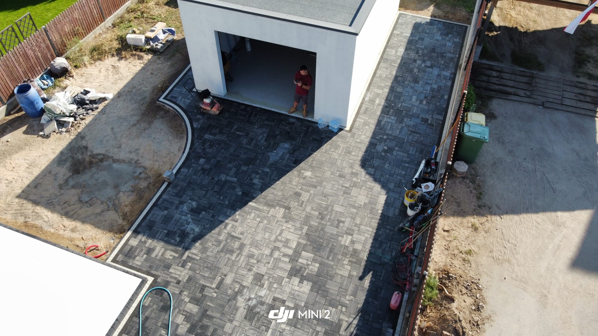 Widok z drona na nowo ułożoną, szarą kostkę brukową przed garażem z otwartymi drzwiami. Na kostce stoi mężczyzna z pilotem. Częściowo wykończony teren po bokach.