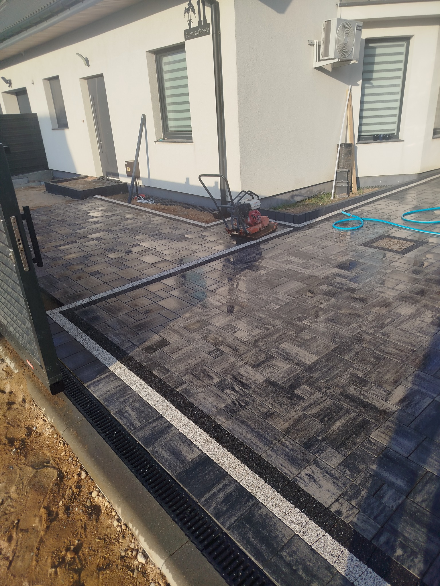 Ułożona kostka brukowa w odcieniach szarości z białymi i czarnymi akcentami. Widoczny drenaż liniowy i zagęszczarka w tle. W tle fragment białego budynku.