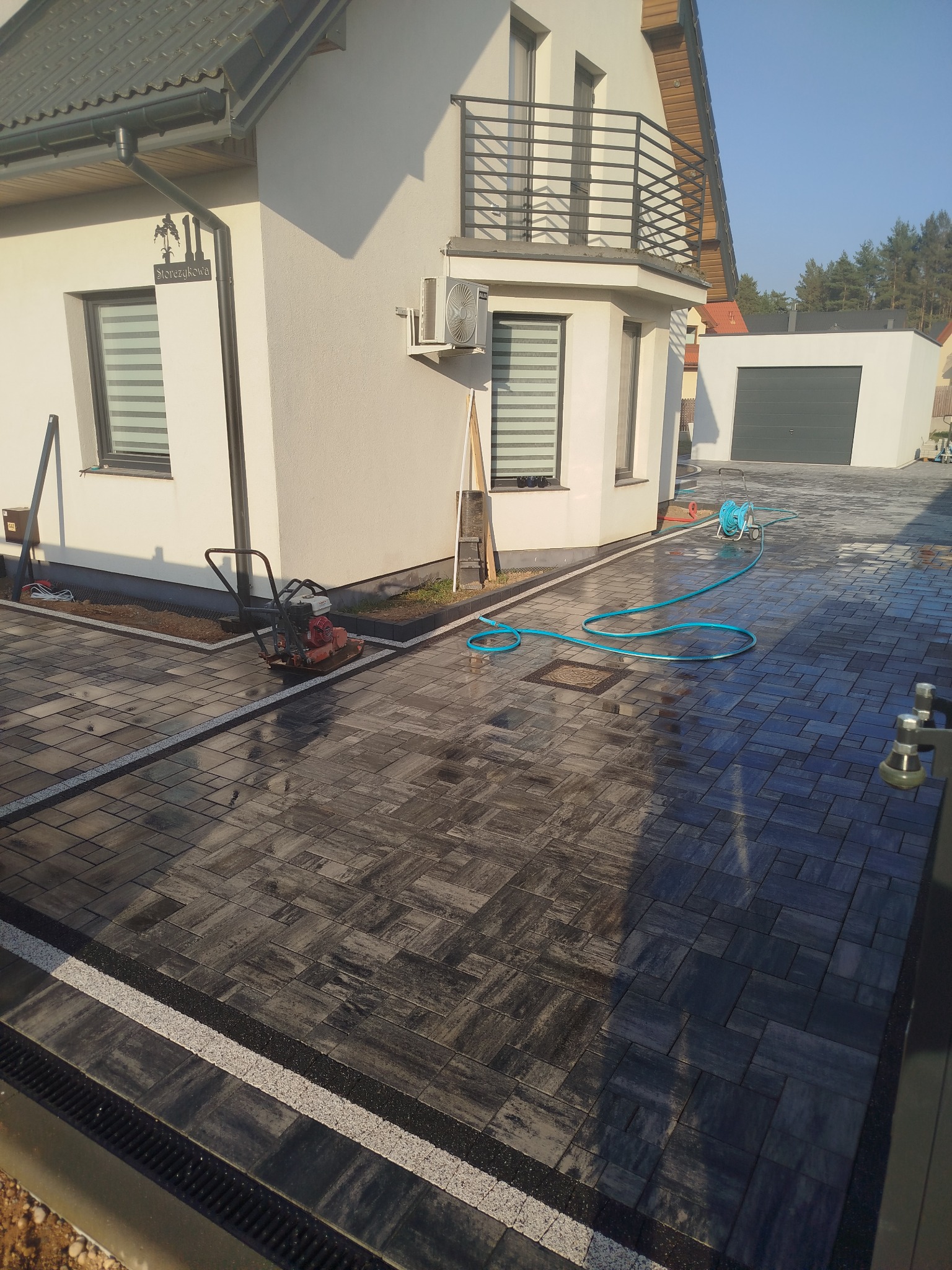 Ułożona kostka brukowa w nowoczesnym wzorze, ciemnoszara z jasnymi akcentami, na tle domu z balkonem i garażem. Widoczny sprzęt budowlany i wąż ogrodowy.