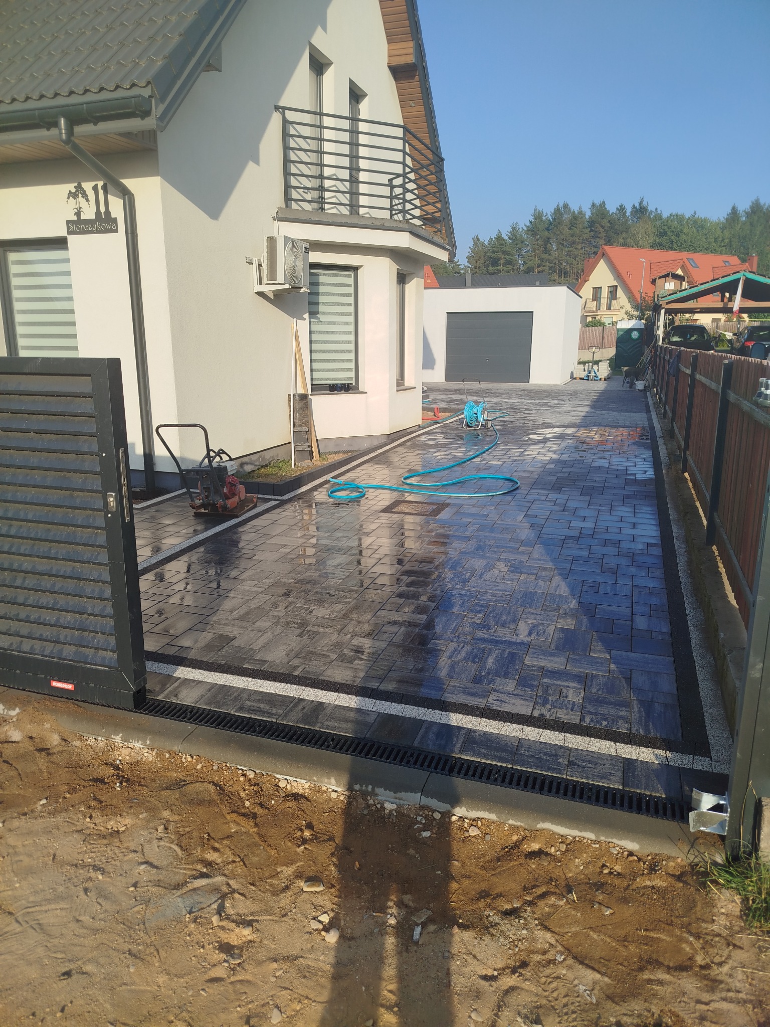 Ułożona kostka brukowa na podjeździe z odpływem liniowym i widoczną zagęszczarką. W tle dom z balkonem i garaż.