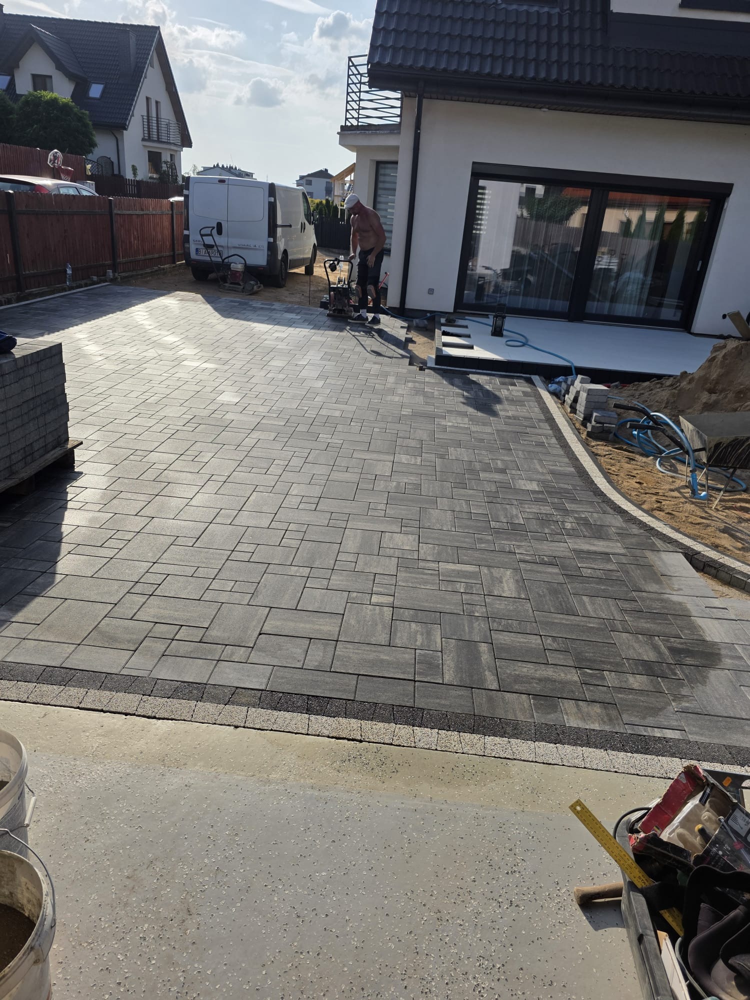 Ułożona kostka brukowa w szarych odcieniach, z obrzeżem z drobnej kostki. Pracownik zagęszcza teren. W tle biały budynek i zaparkowany bus.