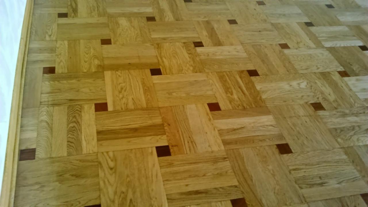 Parkiet ułożony we wzór szachownicy z jasnego drewna z ciemnymi, kwadratowymi intarsjami, widoczna listwa przypodłogowa.