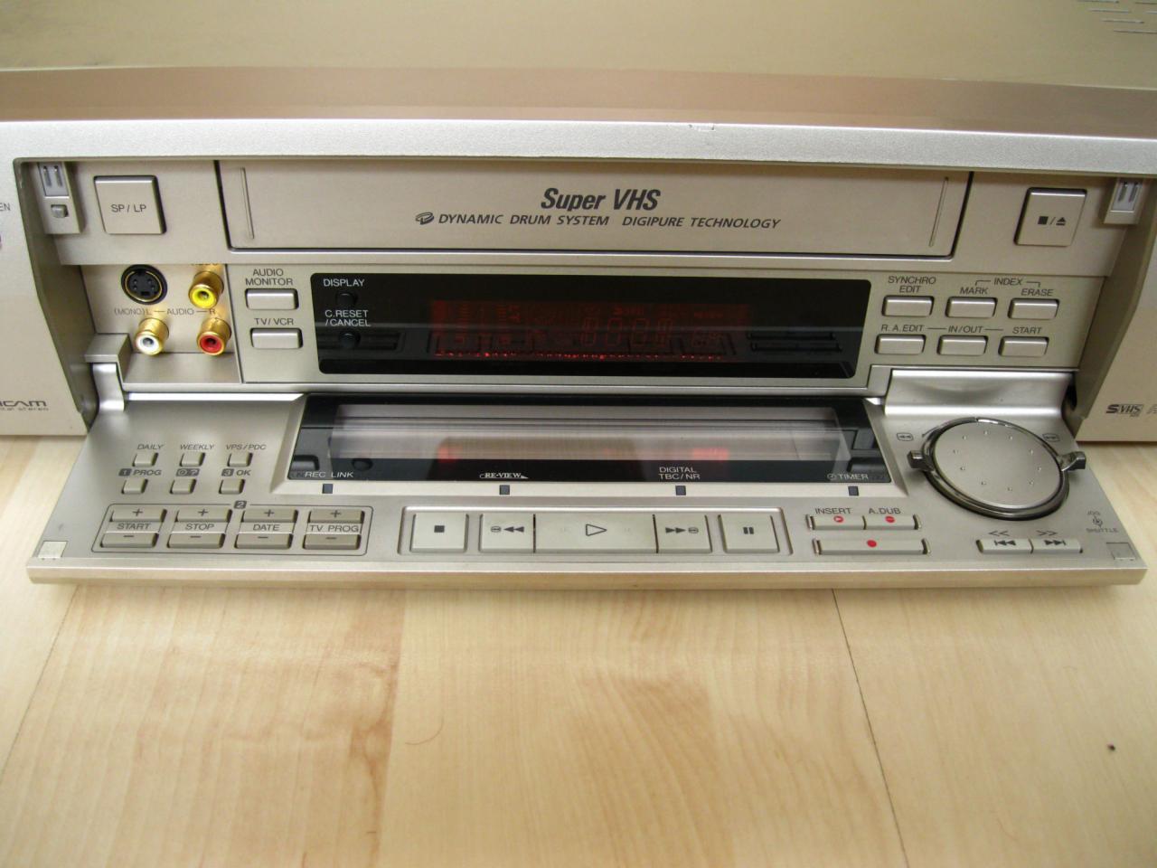 Magnetowid Super VHS w kolorze srebrnym, z widocznymi gniazdami audio i wideo, wyświetlaczem oraz przyciskami sterującymi, leżący na drewnianej podłodze.