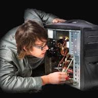 Młody mężczyzna w okularach diagnozuje wnętrze otwartej obudowy komputera stacjonarnego, używając miernika elektrycznego i przyglądając się kablom w słabym oświetleniu.