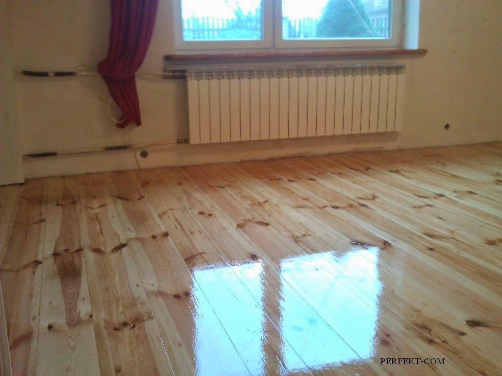 Błyszcząca, świeżo polakierowana drewniana podłoga z widocznym odbiciem okna, grzejnik na ścianie i fragment czerwonej zasłony w rogu.