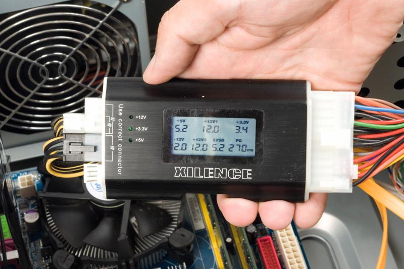 Miernik napięć zasilacza komputerowego marki Xilence, trzymany w dłoni, podłączony do złącza zasilania wewnątrz obudowy komputera stacjonarnego. Wyświetlacz LCD pokazuje wartości napięć +5V, +12V...