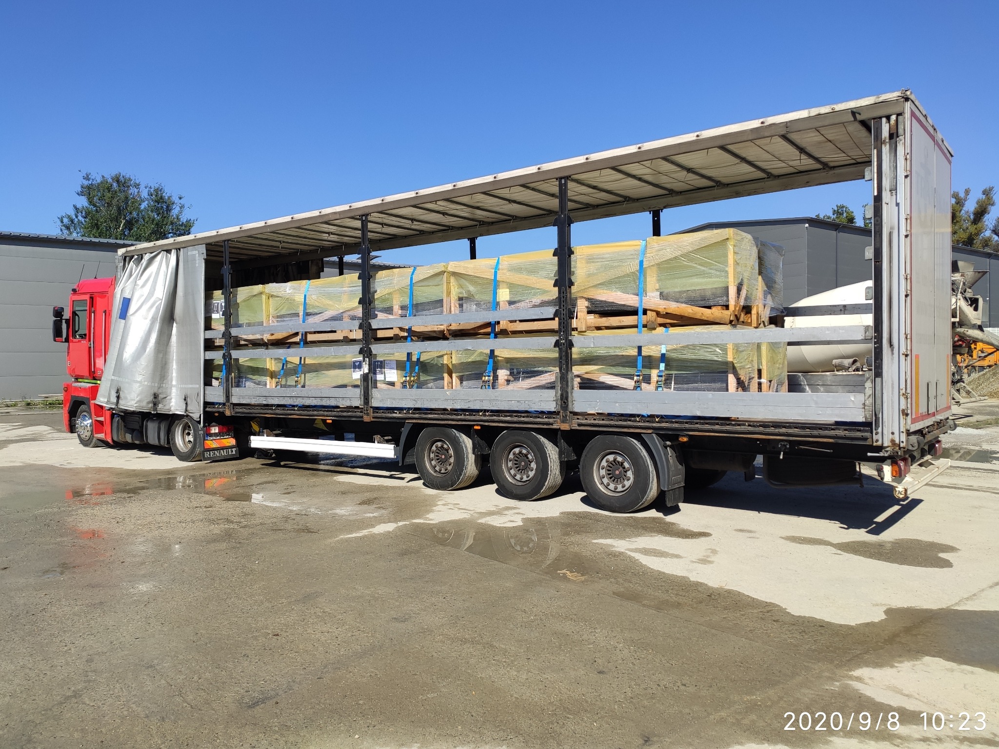 Ciężarówka Renault z naczepą, częściowo załadowana paletami z materiałami budowlanymi, zabezpieczonymi żółtą folią i niebieskimi pasami, na mokrym placu.