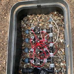 Lewandowski Błażej Green Line - Skrzynka zaworowa z elementami systemu nawadniania, widoczne złączki, elektrozawory i przewody, wypełniona drobnymi kamieniami.