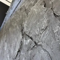 Szara ściana z wyraźną, nieregularną fakturą, imitującą kamień lub skałę, oświetlona z g&oacute;ry, widziana pod kątem. Dekoracyjne wykończenie wnętrza.