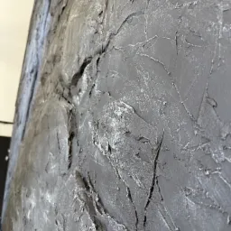 Szara ściana z wyraźną, nieregularną fakturą, przypominającą surowy kamień lub beton, z delikatnymi srebrnymi akcentami, uchwycona pod kątem.