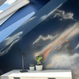 Pokój na poddaszu z oknem dachowym i ścianą ozdobioną malowidłem w stylu chmur. Na biurku lampka i dwie małe rośliny doniczkowe.