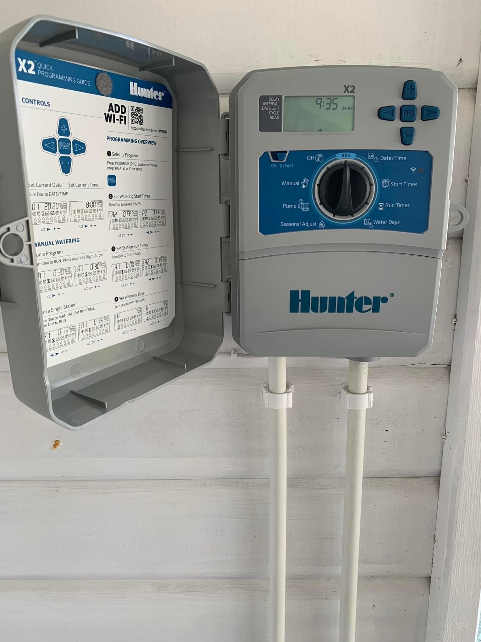 Sterownik nawadniania Hunter X2 zamontowany na ścianie, z otwartą obudową ukazującą panel sterowania, programowanie oraz instrukcję szybkiego programowania.