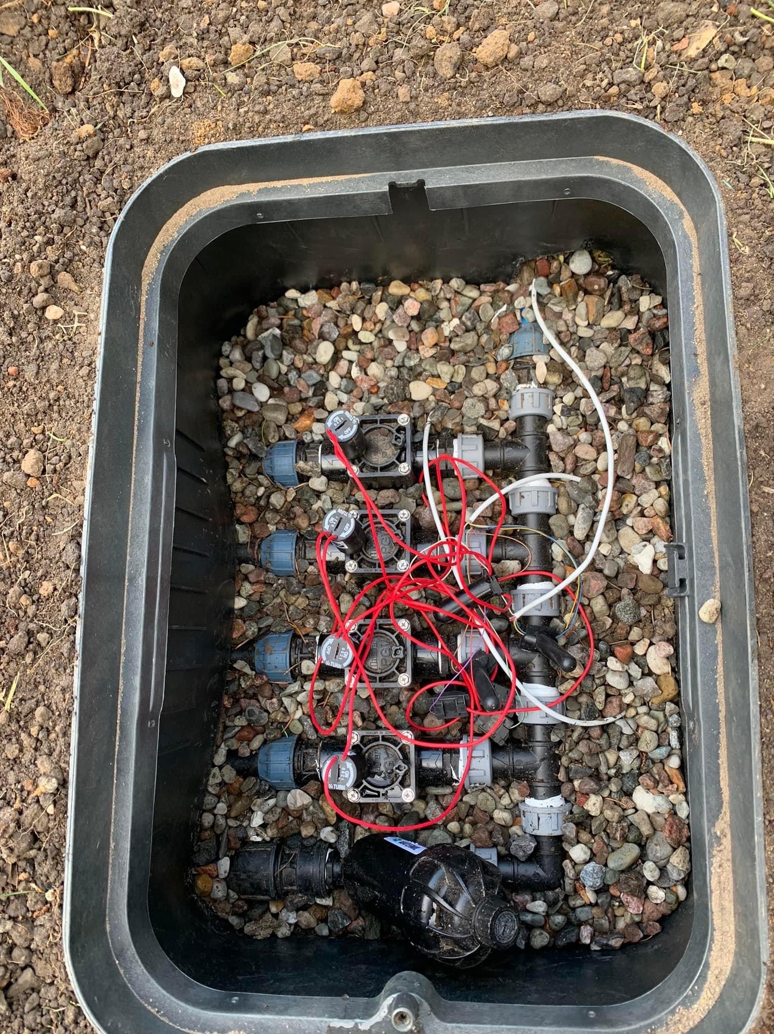 Skrzynka zaworowa z elementami systemu nawadniania, widoczne złączki, elektrozawory i przewody, wypełniona drobnymi kamieniami.