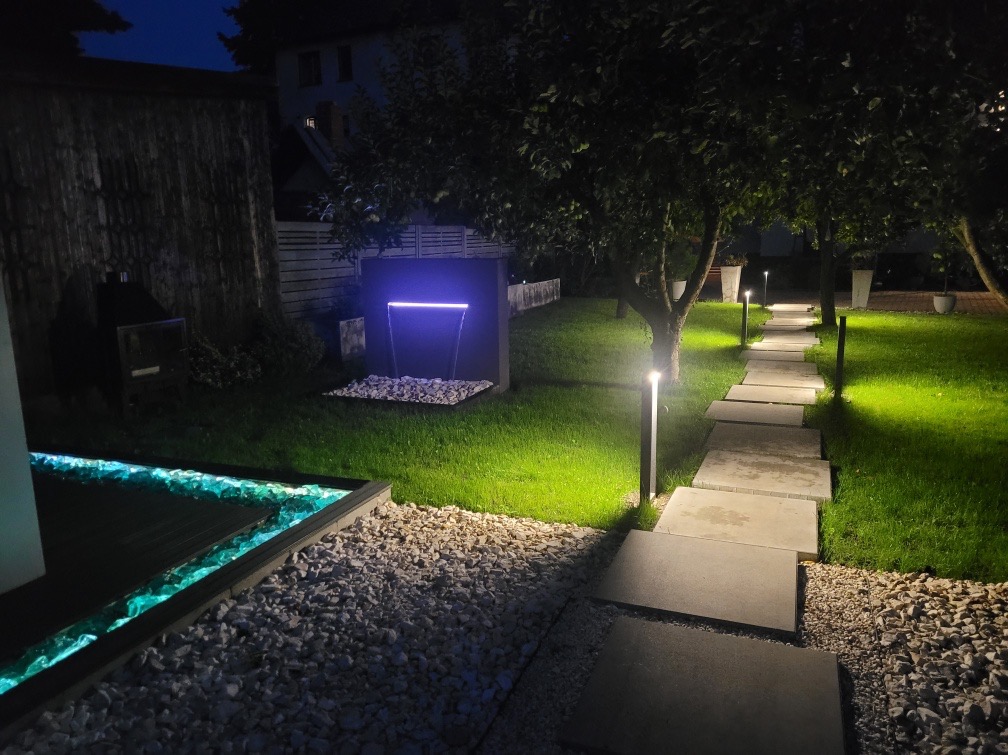 Oświetlony ogród nocą z nowoczesną kaskadą wodną z podświetleniem LED, ścieżką z płyt betonowych i dekoracyjnym żwirem.