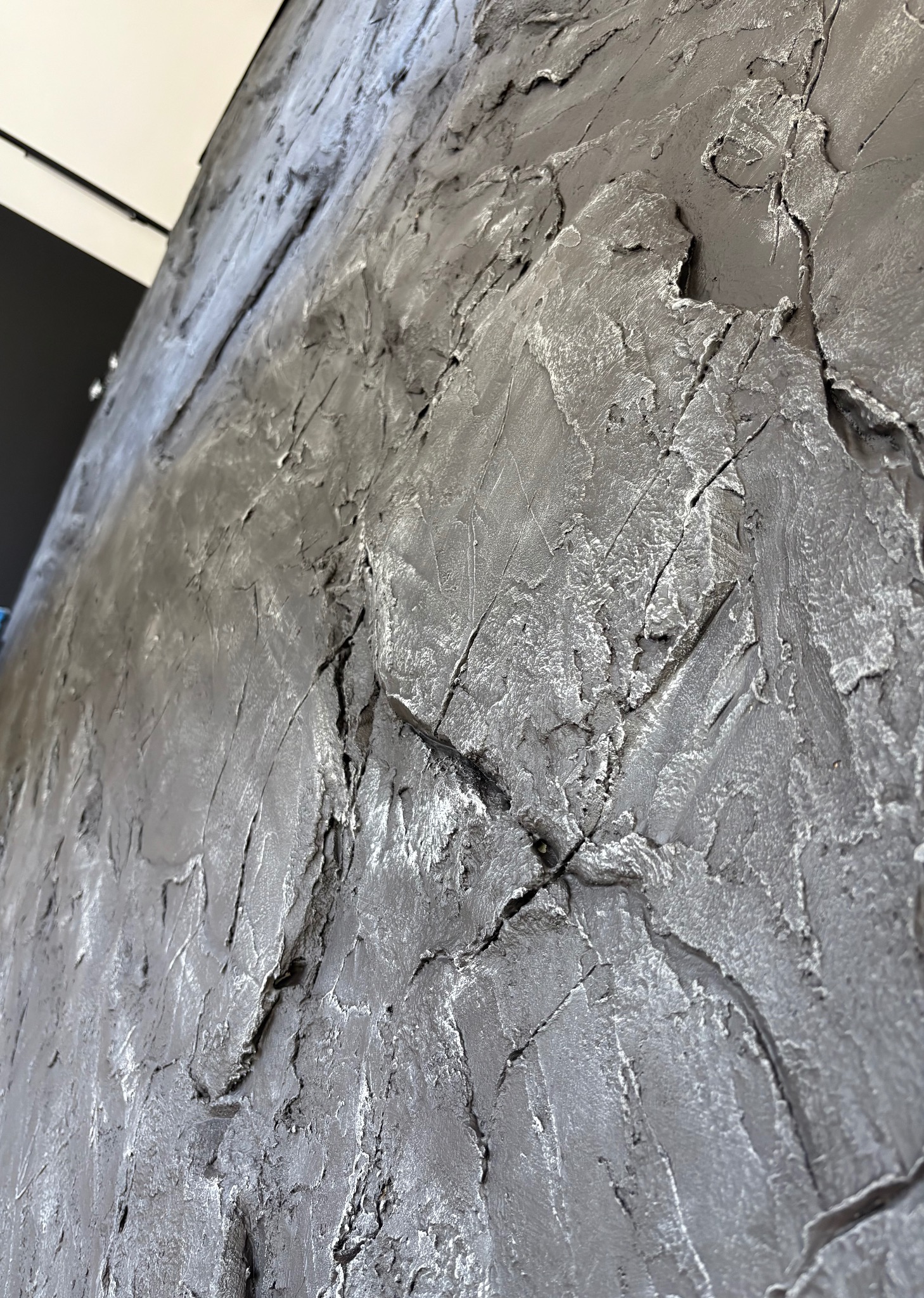 Szara ściana z wyraźną, nieregularną fakturą, imitującą kamień lub skałę, oświetlona z góry, widziana pod kątem. Dekoracyjne wykończenie wnętrza.