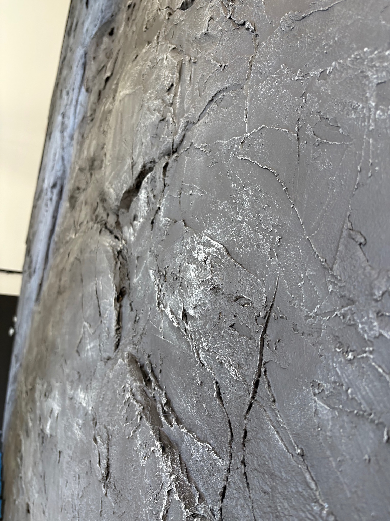 Szara ściana z wyraźną, nieregularną fakturą, przypominającą surowy kamień lub beton, z delikatnymi srebrnymi akcentami, uchwycona pod kątem.