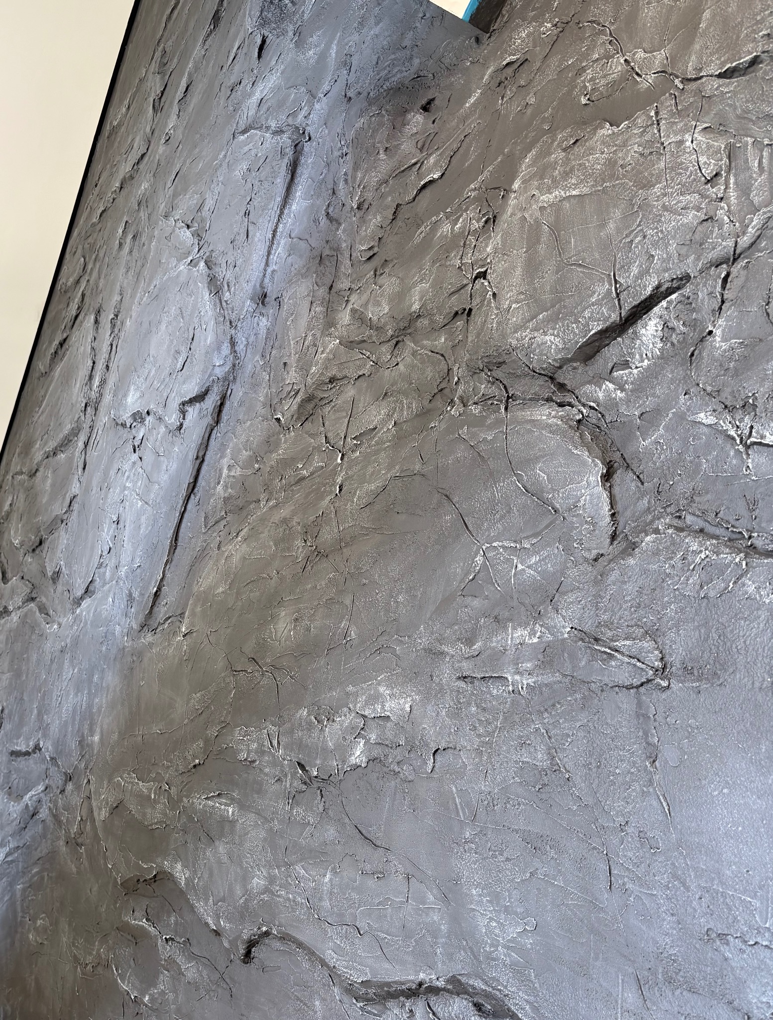 Szara ściana z wyraźną, nieregularną fakturą przypominającą surowy kamień. Widoczne liczne zagłębienia i wypukłości. Oświetlenie podkreśla trójwymiarowość powierzchni.