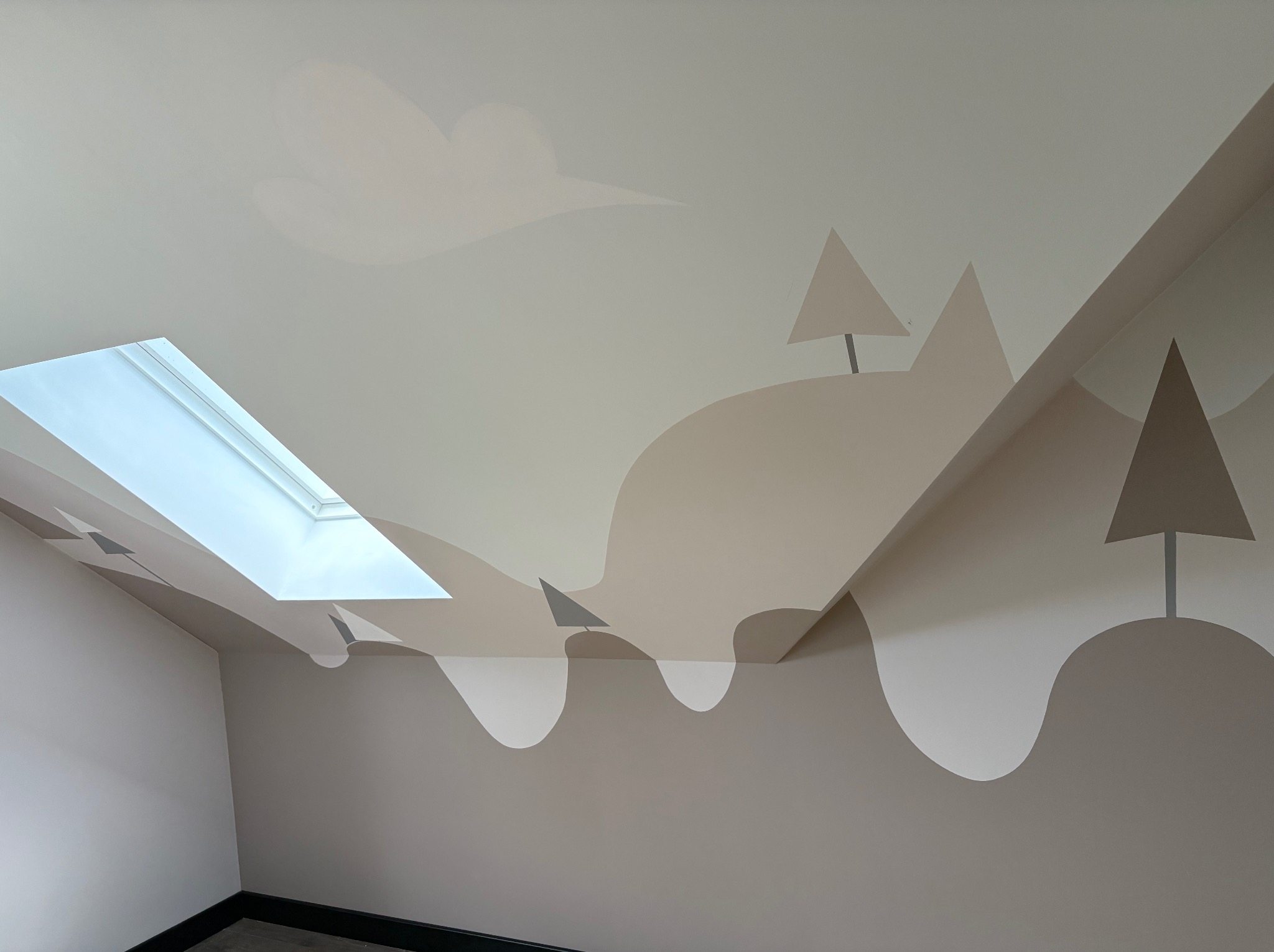 Pokój na poddaszu z oknem dachowym i ręcznie malowaną, dziecięcą grafiką na ścianie, przedstawiającą wzgórza i drzewa w odcieniach beżu i szarości.