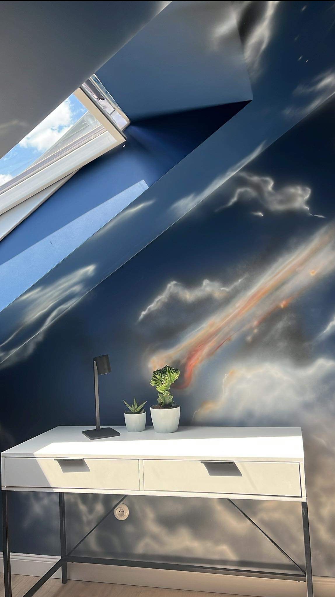Pokój na poddaszu z oknem dachowym i ścianą ozdobioną malowidłem w stylu chmur. Na biurku lampka i dwie małe rośliny doniczkowe.