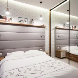 Nowoczesna sypialnia z łóżkiem z szarym, tapicerowanym zagłówkiem z drewnianą półką, szafką nocną i lustrzaną szafą przesuwną.