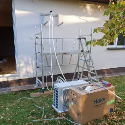 Montaż klimatyzacji: jednostka zewnętrzna Haier, rusztowanie aluminiowe, przewody i uchwyt na ścianie budynku jednorodzinnego w trakcie instalacji.