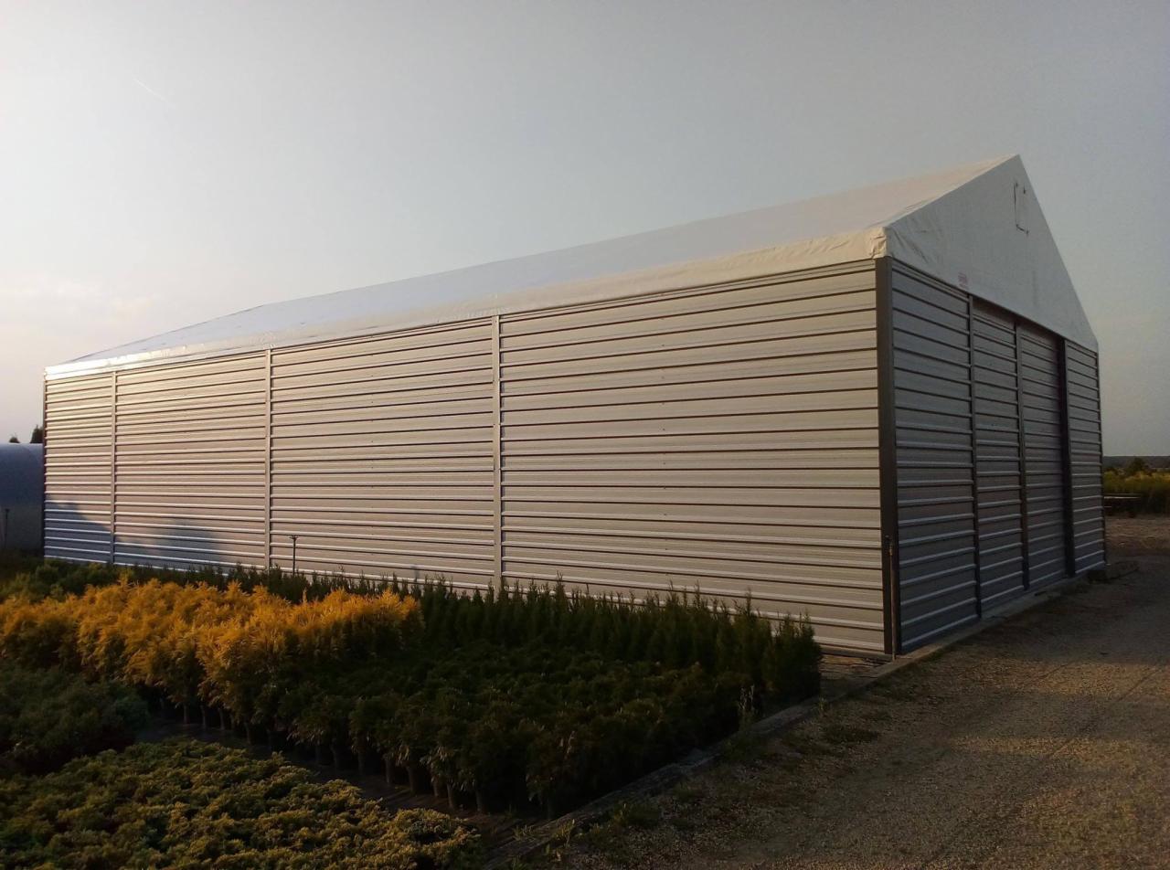 Gotowa hala z blachy trapezowej z białym dachem plandekowym, widok z boku na tle upraw szkółkarskich.