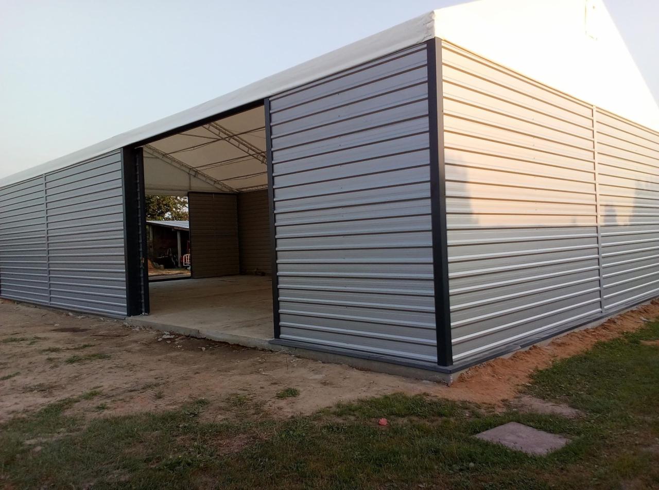 Gotowa hala z blachy trapezowej w kolorze szarym z otwartymi przesuwnymi drzwiami, widok z zewnątrz na betonową posadzkę wewnątrz.