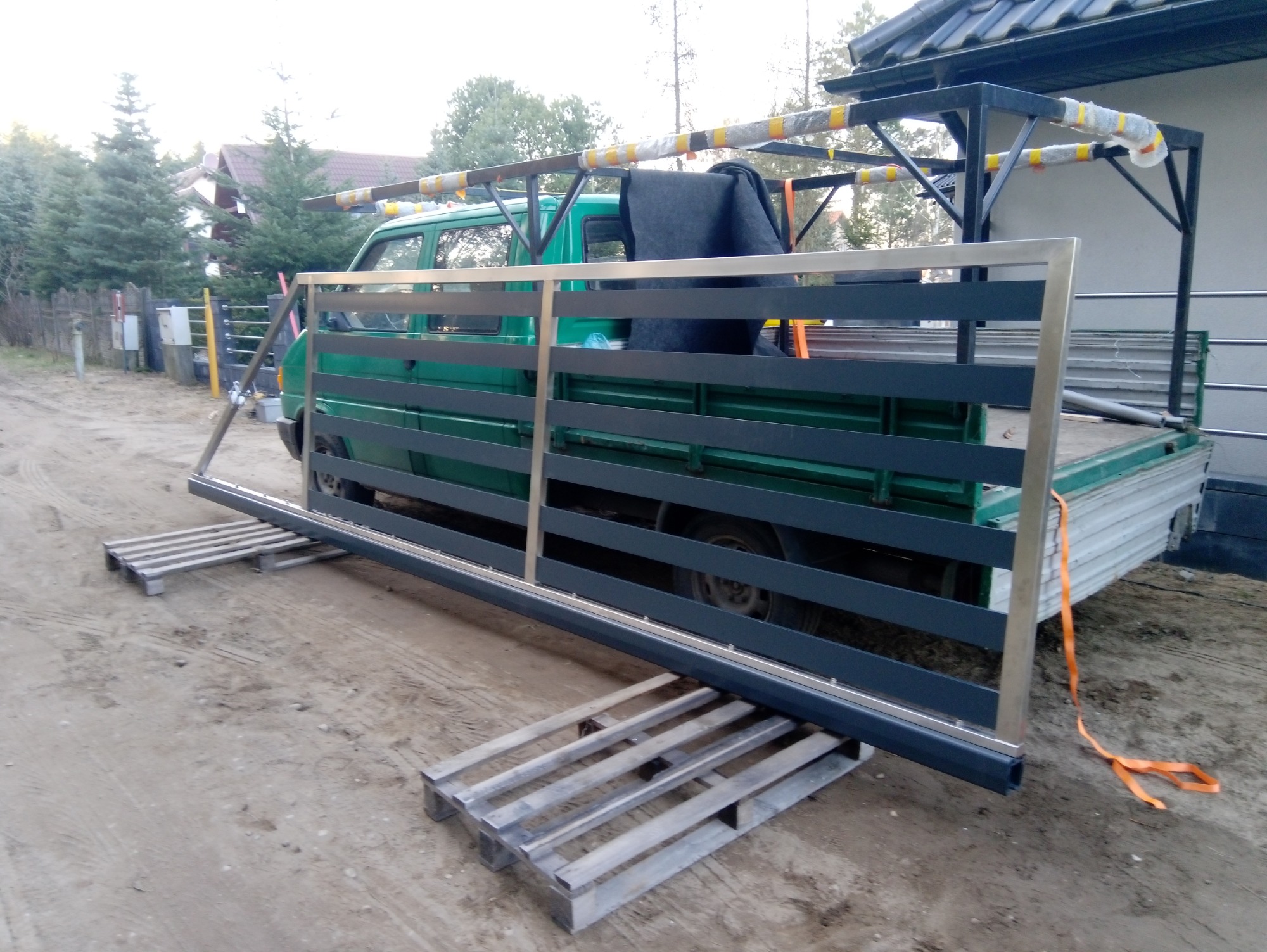 Częściowo zmontowane przęsło ogrodzenia metalowego, oparte na paletach, przewożone na pace zielonego samochodu dostawczego z konstrukcją wspornikową na dachu, na tle piaszczystego podłoża i zabudowań.