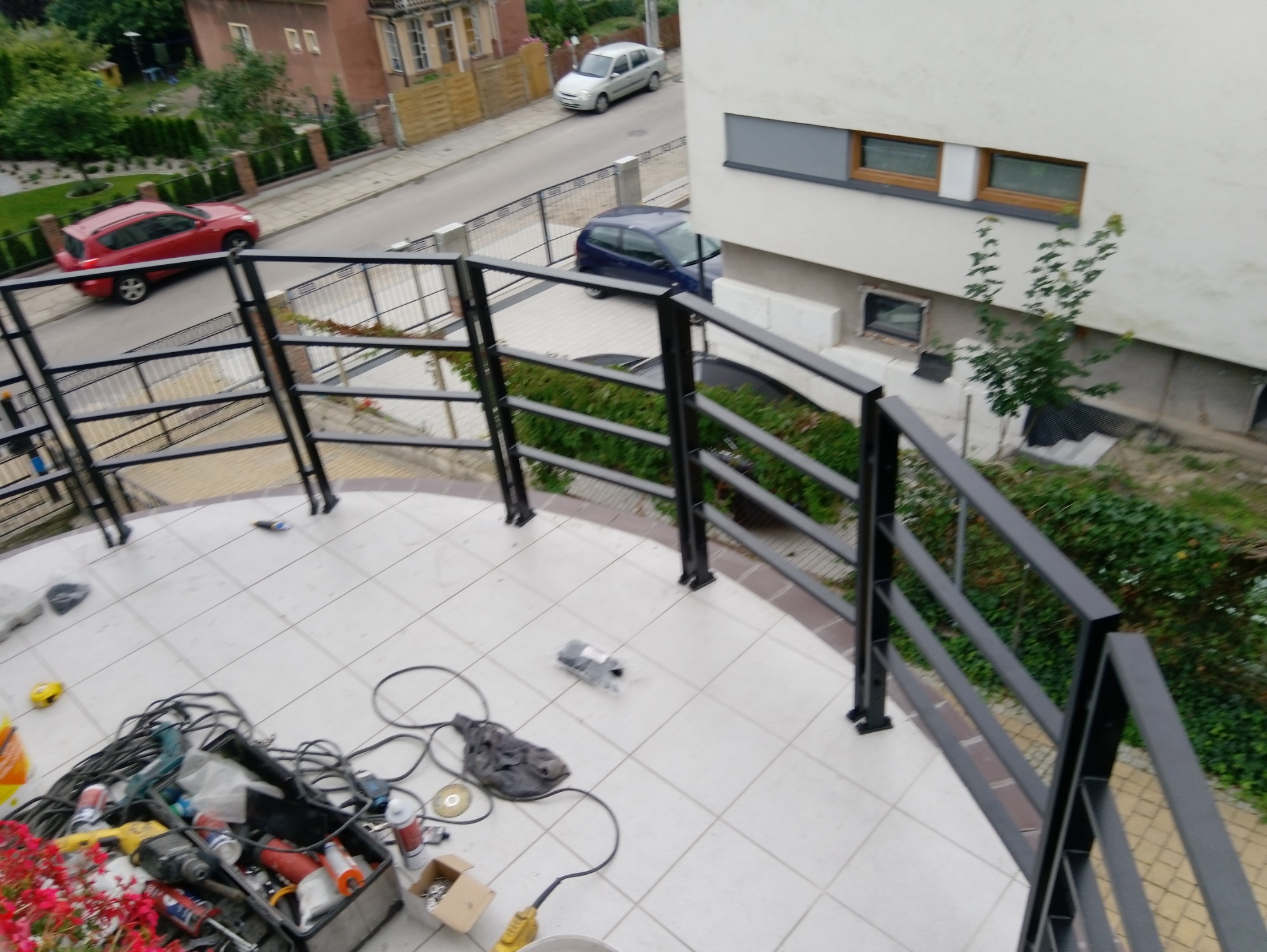 Czarna, metalowa balustrada na balkonie z białymi płytkami, widoczne narzędzia i skrzynka z akcesoriami, ujęcie z góry na tle ulicy z samochodami.
