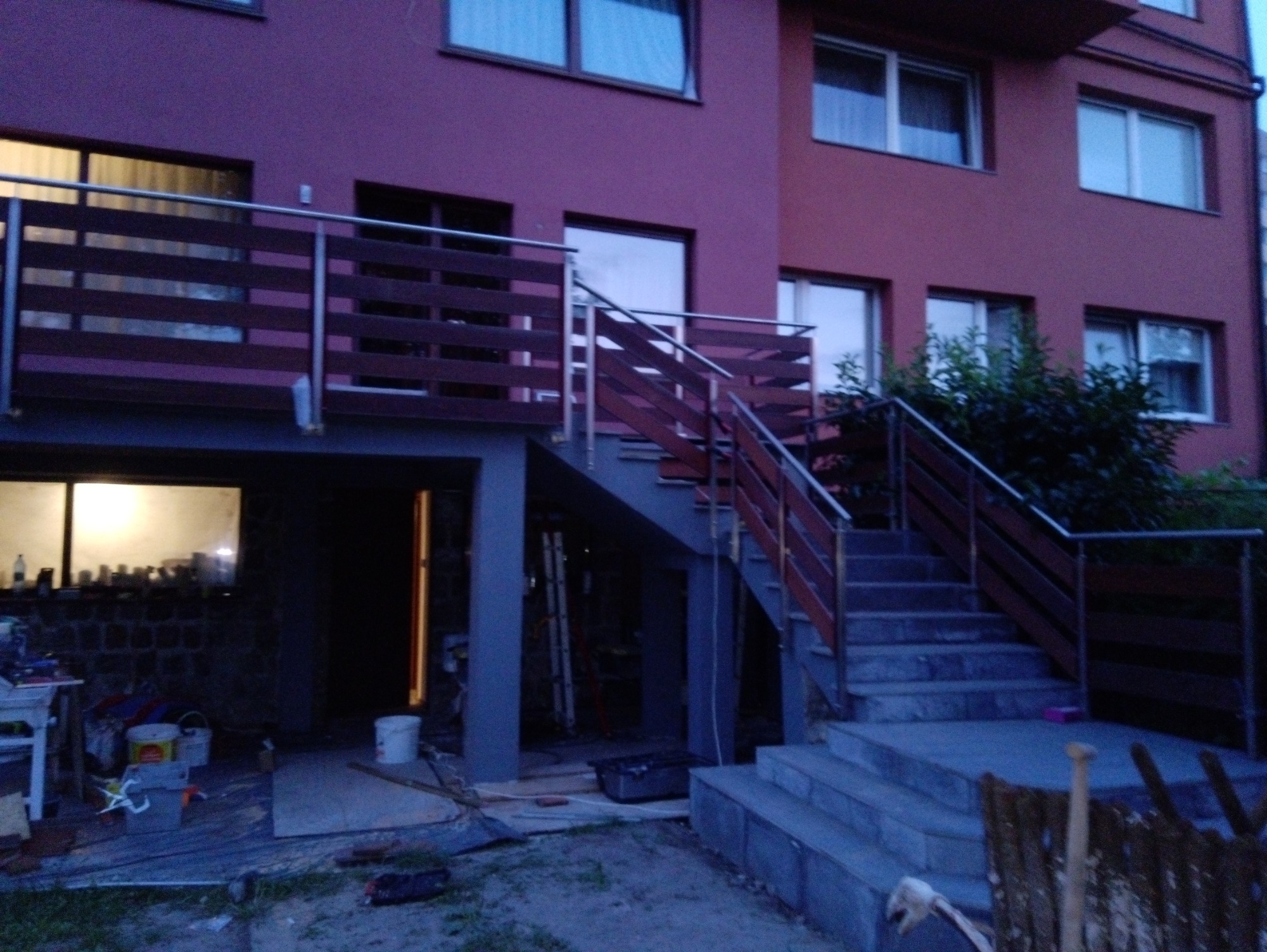 Dwukondygnacyjny budynek z balkonem i schodami zewnętrznymi, zabezpieczonymi drewnianymi balustradami z metalowymi poręczami, widoczne materiały budowlane i narzędzia.