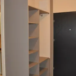 Szara szafa wnękowa z otwartymi półkami i drążkiem na ubrania, zamontowana przy ścianie w przedpokoju obok czarnych drzwi wejściowych.