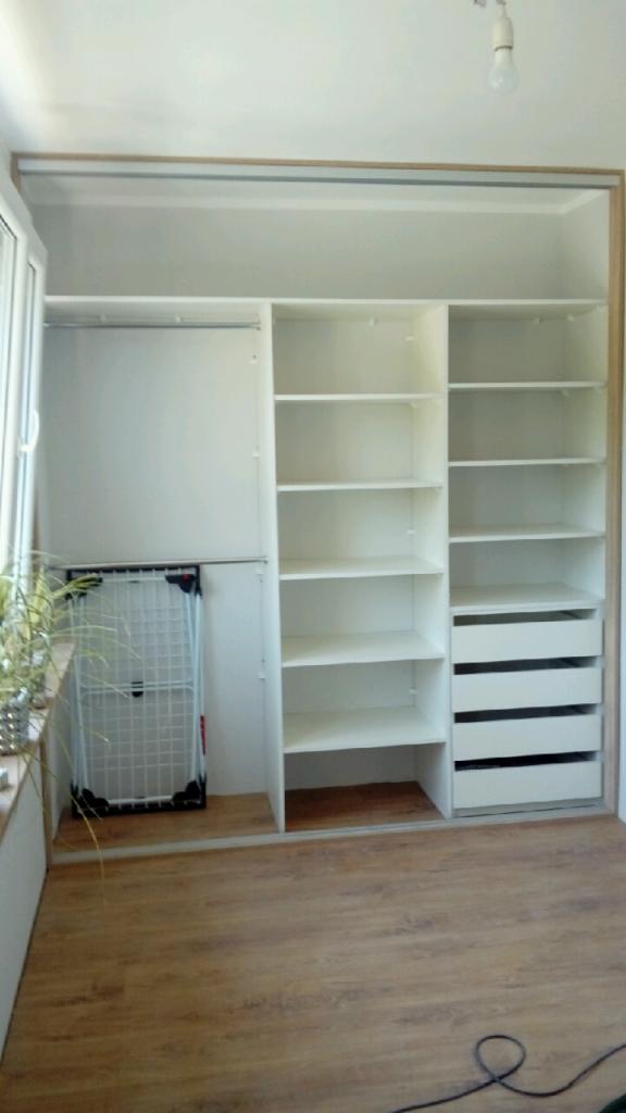 Biała szafa wnękowa z otwartymi półkami i szufladami, widoczna suszarka na pranie i roślina doniczkowa przy oknie, podłoga z paneli.