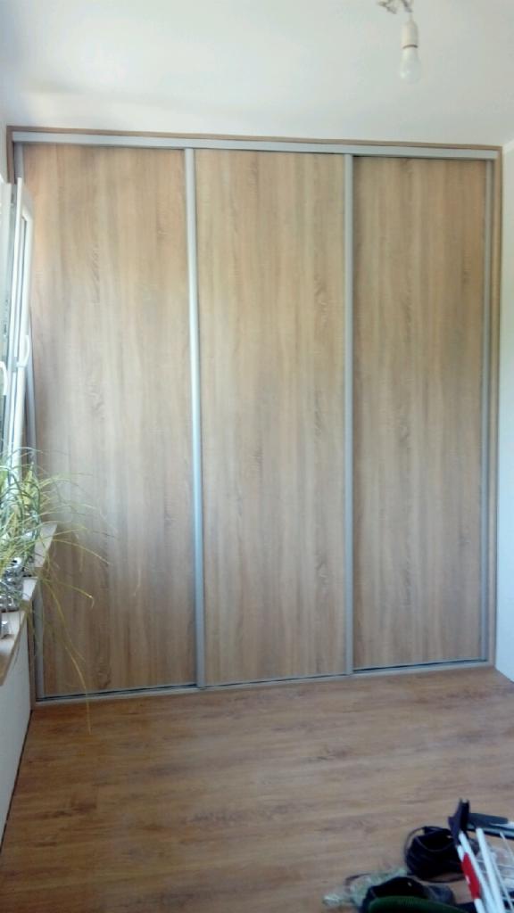 Trzydrzwiowa szafa wnękowa z przesuwnymi drzwiami o drewnianym wzorze i srebrnych obramowaniach, widoczna na tle drewnianej podłogi w jasnym pomieszczeniu.