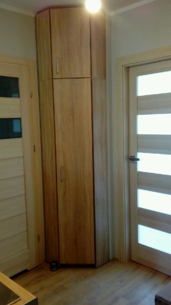 Wysoka, drewniana szafa wnękowa z dwoma drzwiami, dopasowana do narożnika pomieszczenia między dwoma drzwiami wejściowymi z przeszkleniami.