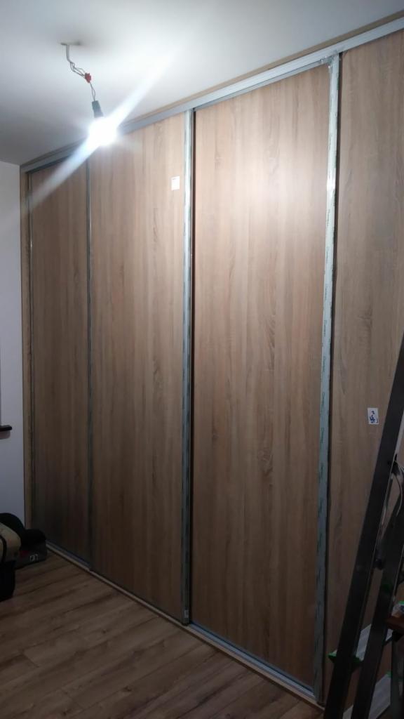 Zamknięta szafa wnękowa z przesuwnymi drzwiami o drewnopodobnym wykończeniu, widoczna drabina oparta o bok szafy i prowizoryczne oświetlenie sufitowe.