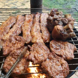 Szeroki kadr na grill z różnymi rodzajami mięsa: długimi paskami boczku, stekami i golonką, z widocznym ogniem pod rusztem i szczypcami do obracania mięsa.