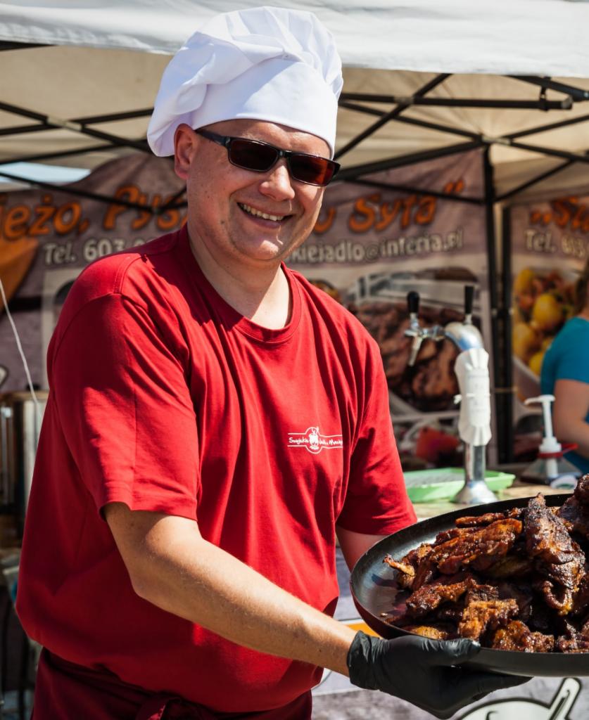 Uśmiechnięty kucharz w okularach przeciwsłonecznych i czapce kucharskiej prezentuje tacę z pieczonym mięsem na tle stoiska z napisem i dystrybutorem piwa.