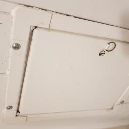 Sanpa Sp. z o.o. - Biała klapa rewizyjna z metalowym uchwytem w ramie przymocowanej śrubami do ściany, widoczne ślady zabrudzeń i uszkodzeń na powierzchni.