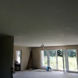 Surowe wnętrze pokoju z gładzią na ścianach i suficie, duże okna balkonowe z widokiem na zieleń, widoczne narzędzia i materiały budowlane, przygotowane do dalszych prac wykończeniowych.