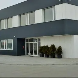 Nowoczesny budynek biurowy z szarymi ścianami i białymi panelami, widoczne okna PCV, zaparkowane samochody i małe drzewka przed wejściem.