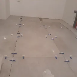 Przygotowanie do układania płytek podłogowych w Pruszkowie, widoczne krzyżyki dystansowe i poziomowanie. W tle instalacja wodno-kanalizacyjna.