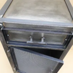 Metalowa szafka nocna z szufladą i ażurowymi drzwiczkami, spawana z profili stalowych, widoczne spawy i surowe wykończenie.