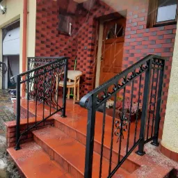 Czarna, metalowa poręcz z ozdobnymi elementami na schodach wejściowych do domu z czerwoną cegłą; osoba w oddali czyści podjazd myjką ciśnieniową w zimowy dzień.